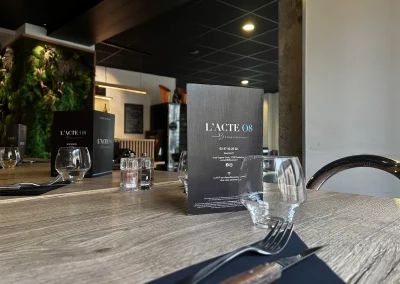 Gros plan sur une table dressée au restaurant L’Acte 08, mettant en avant la carte de la brasserie, des verres raffinés et un mur végétal en arrière-plan.