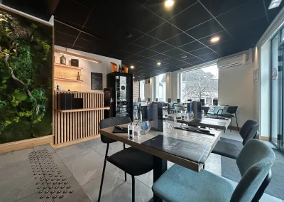 Vue de la salle du restaurant L’Acte 08, mettant en avant des tables dressées, un mur végétal, une cave à vin et de grandes baies vitrées pour un cadre lumineux.