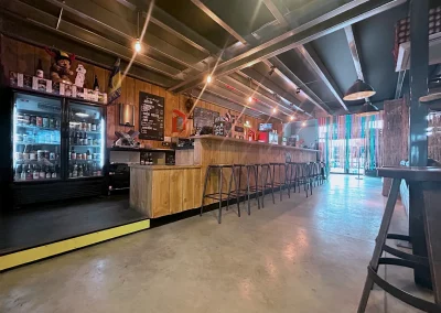 Vue en perspective du bar en bois massif de Beer Or Not To Beer, avec ses tabourets métalliques, un éclairage chaleureux et une ambiance industrielle authentique.
