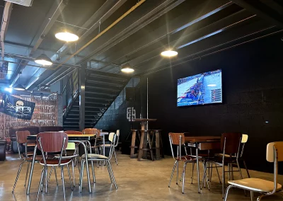 Espace détente avec tables et chaises rétro, ambiance industrielle, télévision murale diffusant un programme sportif et escalier menant à la mezzanine.