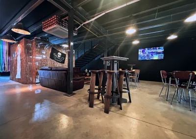 Vue de l’espace détente avec tables hautes en bois, canapés en cuir, TV murale diffusant un événement sportif et, sur la gauche, l’espace cave à bière à la vente.