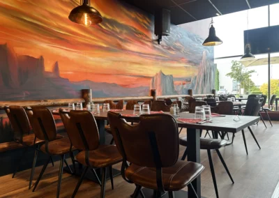 Salle du Dakota Café avec fauteuils en cuir brun, tables dressées et fresque panoramique représentant un coucher de soleil sur le désert.