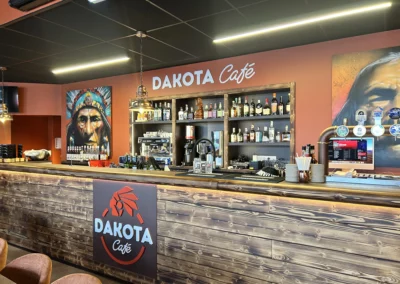 Bar du Dakota Café avec comptoir en bois, enseigne murale, étagères à bouteilles et suspensions métalliques.